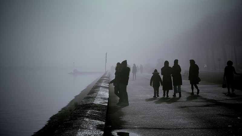 Dark silhouettes of people walking along the lake on a background of fog.