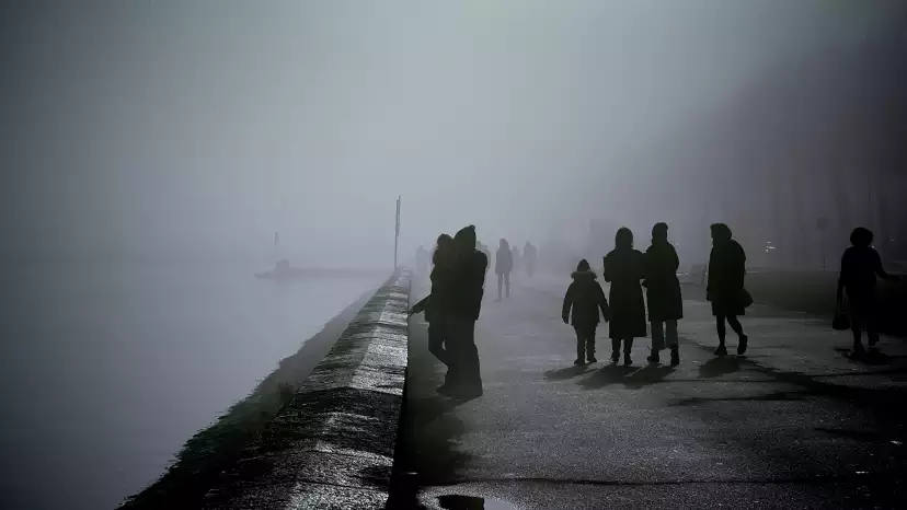 Dark silhouettes of people walking along the lake on a background of fog.