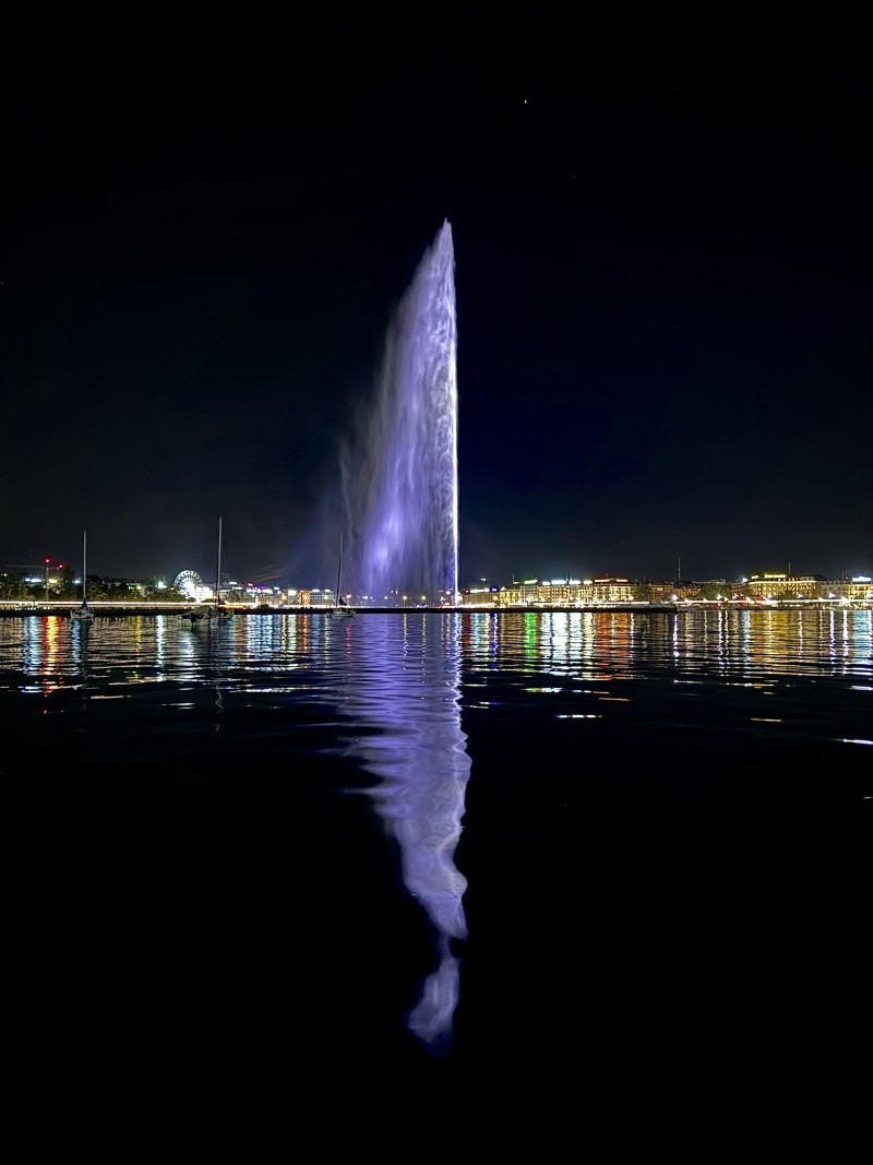 Geneva Jet d'Eau illuminated purple at night reflecting into the harbour with the city lights behind