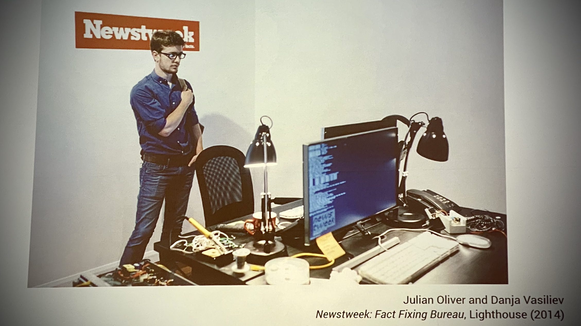 A man behind a desk with Newstweek logo on the wall