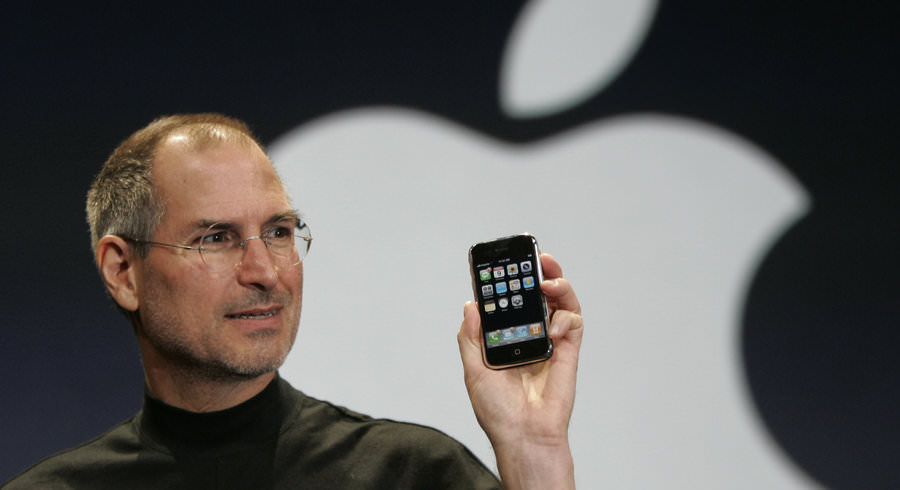 A close-up of Steve Jobs holding the original iPhone in his left hand with a glowing Apple logo behind