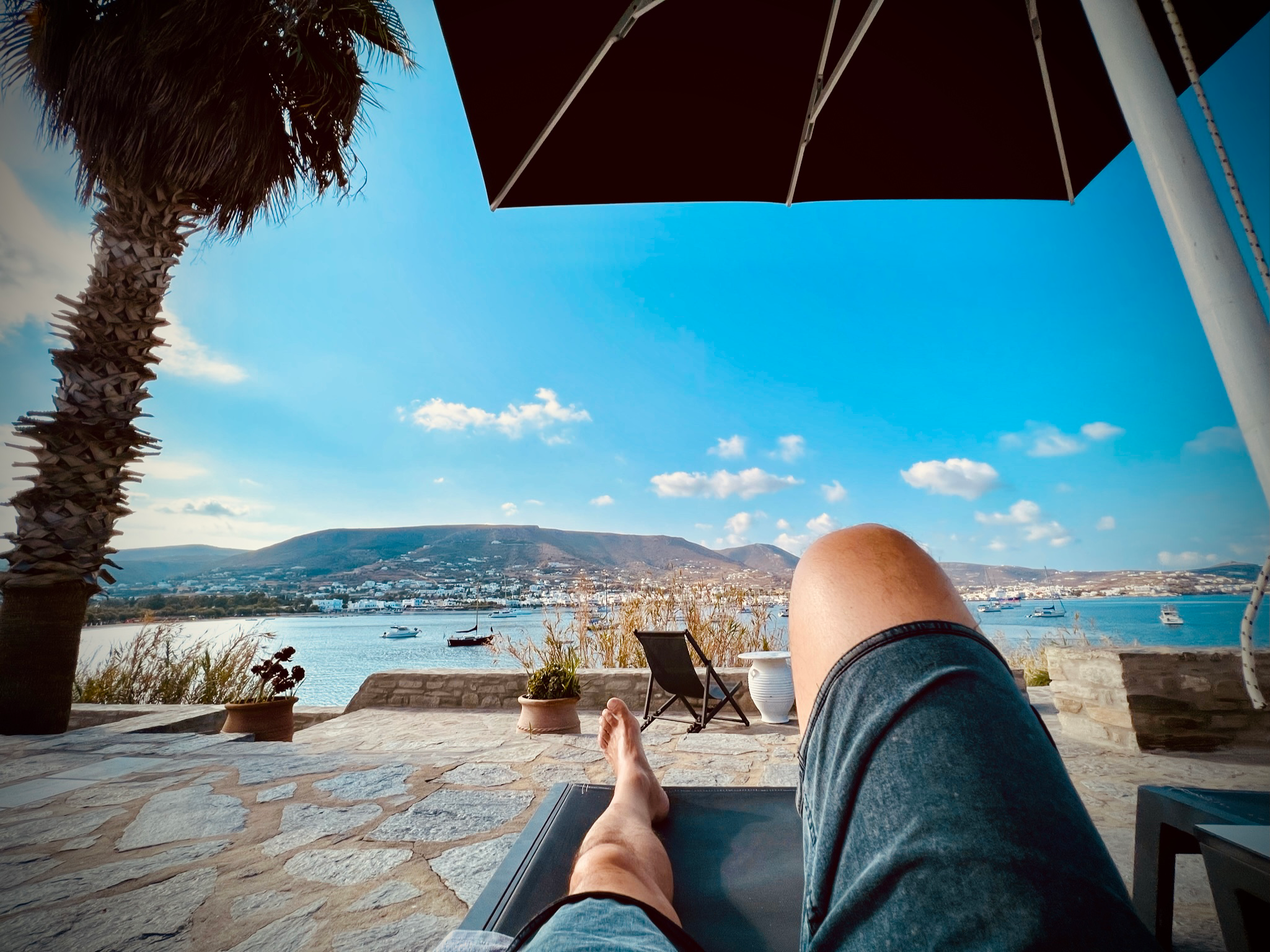 A view of Parikia bay from the Parian Village Hotel laying on a deck chair under a parasol with dotted blue sky as backdrop